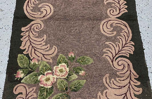 A close-up of a woven rug or textile with a dark brown center, beige scroll motifs along the sides, and pink-and-green floral clusters in the lower left corner.