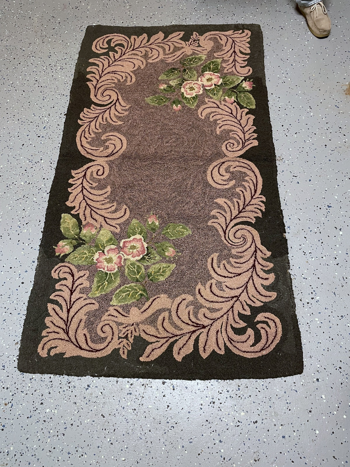 A rectangular vintage-style area rug with a dark brown center and a raised beige scrollwork border, decorated with pink roses and green leaves near the top and bottom corners, shown on a speckled gray floor.