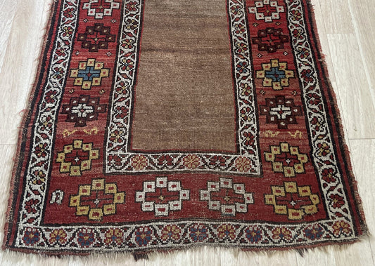 Close-up of a 150-year-old hand-knotted Kurdish rug showing the rich camel/tobacco field, a crimson main border with varied geometric motifs in ivory, saffron gold, dark brown, and teal blue, plus crisp ivory guard borders with vine-and-leaf scrolls.