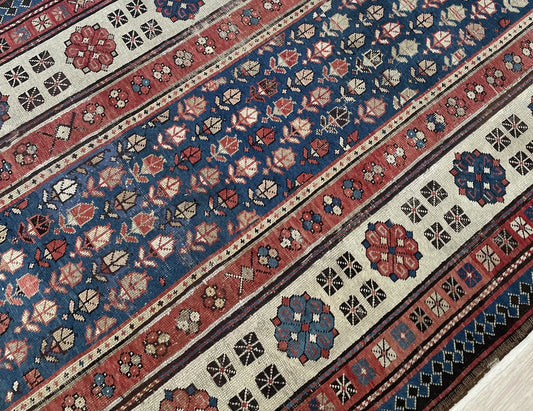 Close-up of the corner of an antique Caucasian Talish carpet showing the transition from the indigo lattice field to the wide ivory main border with octagonal medallions and the teal outer diamond border.


