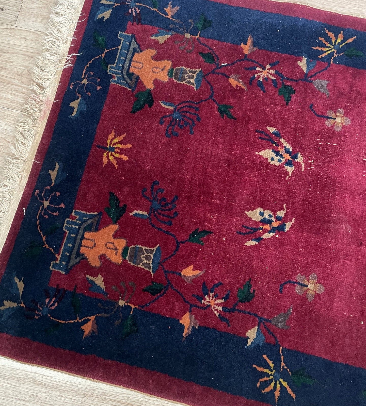 Close-up of the corner of an antique Chinese Art Deco rug showing the transition from the open crimson field with butterflies to the midnight navy border with bronze vessel motifs.

