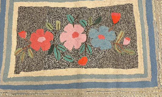 Angled rug corner showing black edging, pale textured border strips and a coral flower.