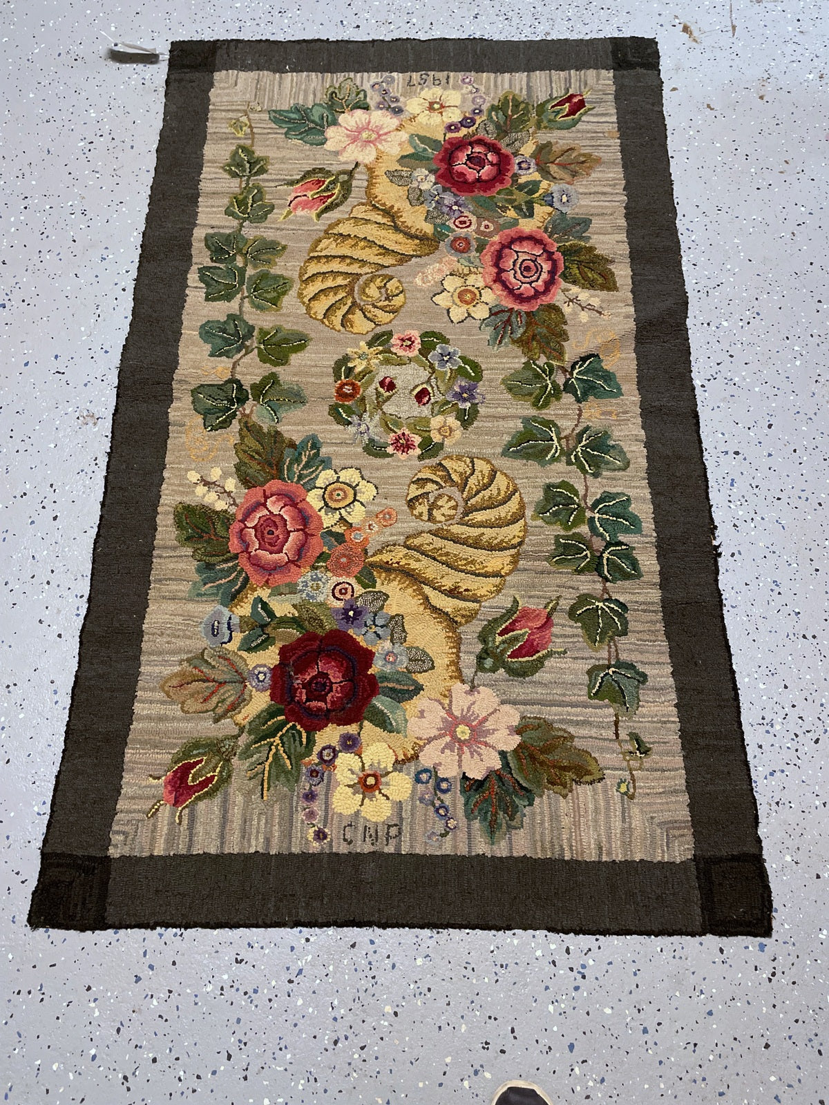 Angled rug end with burgundy flowers, a curling golden horn and hooked initials.