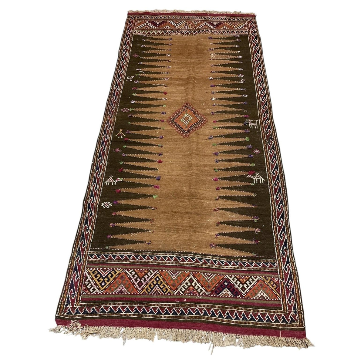 Full view of a 1930s wool kilim with camel centre, dark serrated panels, central diamond and pale fringes.