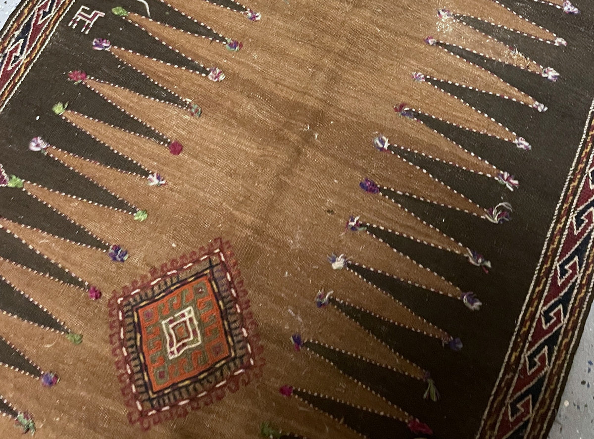 Central diamond and stylised figures among pointed brown panels accented with colourful yarn tufts.