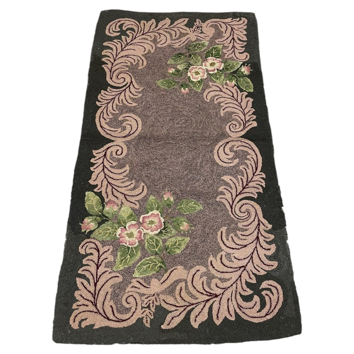A rectangular vintage-style rug with a dark charcoal gray background, featuring a raised beige scrollwork border and pink and green floral motifs near the top and bottom corners.