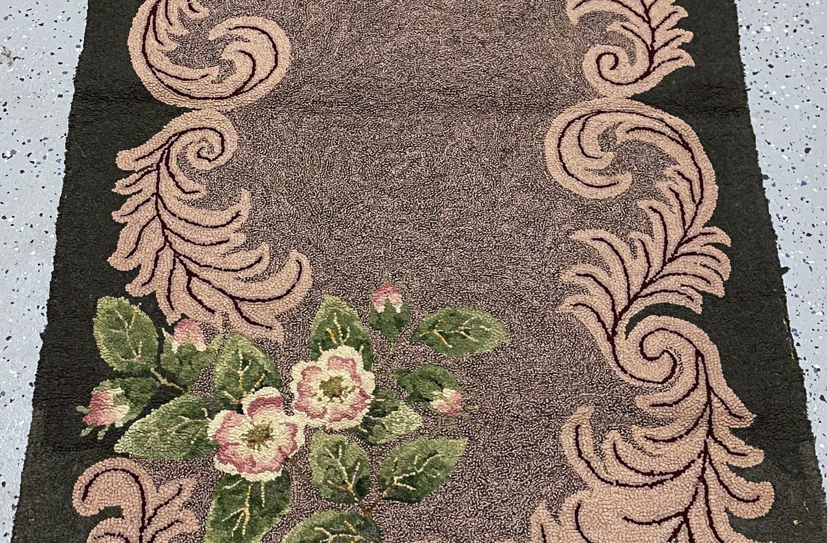A close-up of a woven rug or textile with a dark brown center, beige scroll motifs along the sides, and pink-and-green floral clusters in the lower left corner.