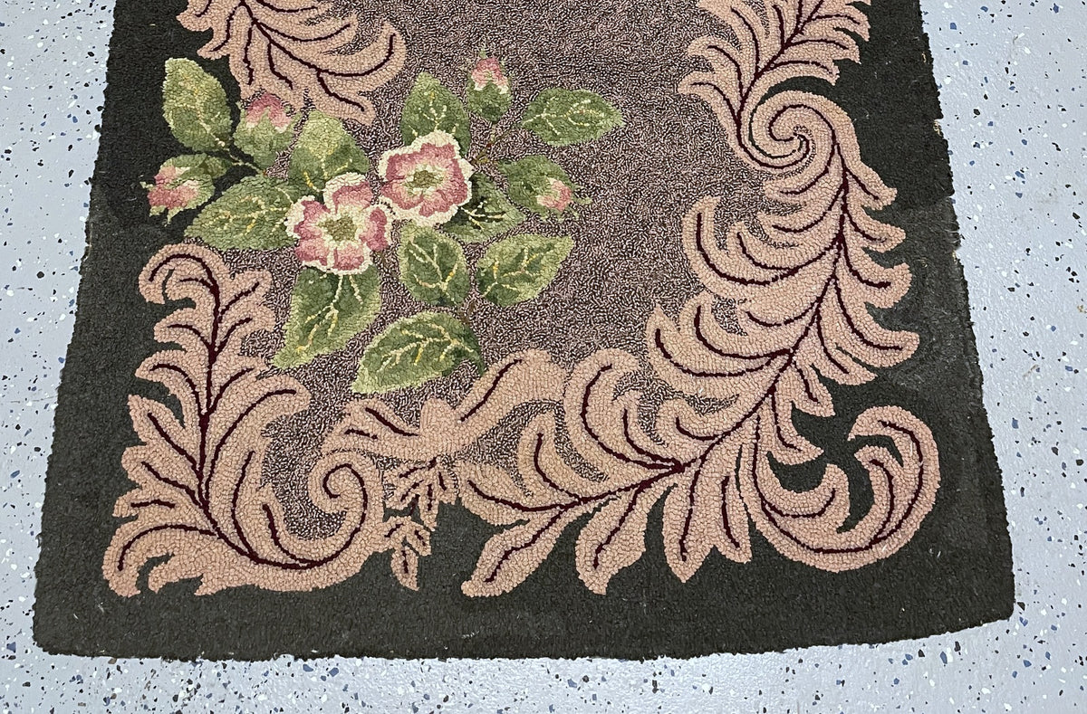 A square vintage-style rug with a dark charcoal background, featuring a central cluster of pink flowers and green leaves surrounded by a raised beige scrollwork border.