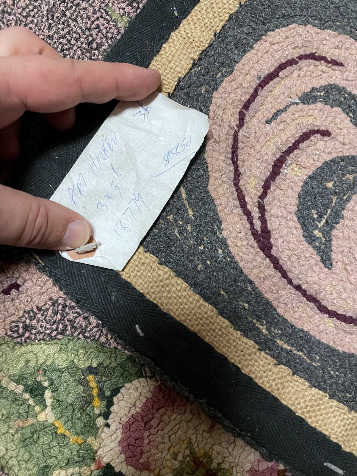A close-up of a hand holding a small white paper tag with handwritten text over a patterned textile rug. The rug has a dark gray and beige striped border with a pink and dark gray spiral motif.