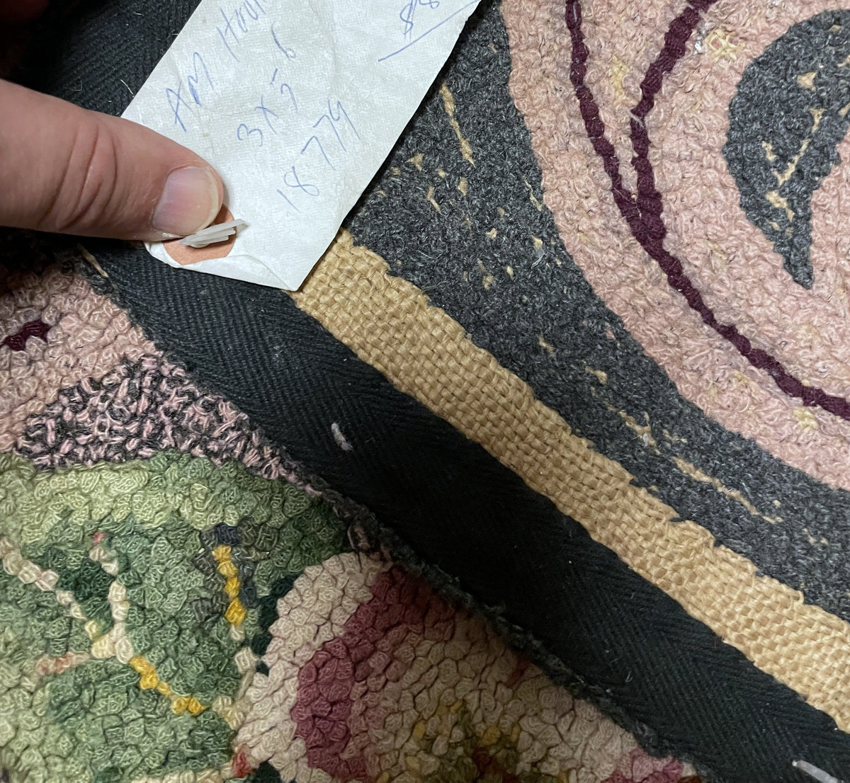 A close-up of a patterned textile with a hand pointing to a small white tag attached near the edge. The fabric has a multicolor floral and abstract design in muted pink, green, black, beige, and burgundy tones.