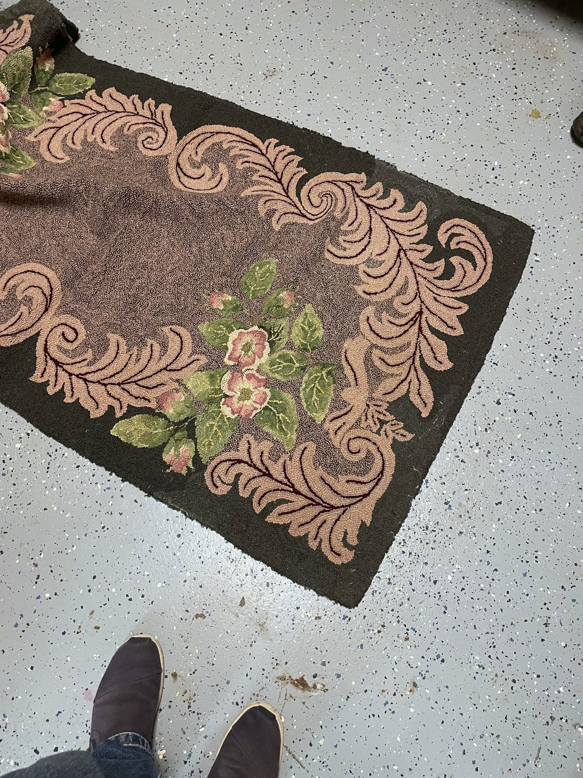 A rectangular vintage-style area rug with a brown center, a beige scroll border, and pink floral accents, shown on a speckled gray floor with a person's feet visible at the bottom.