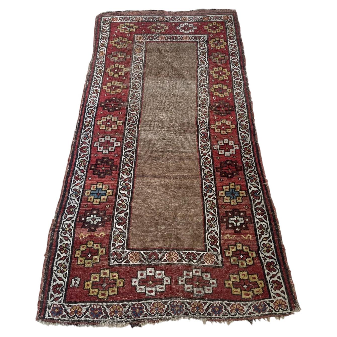Full view of an antique Kurdish tribal wool rug from the 1870s featuring an open undecorated field in warm natural undyed camel wool, framed by a wide deep crimson border with bold geometric medallions including hooked diamonds and eight-pointed star rosettes.

