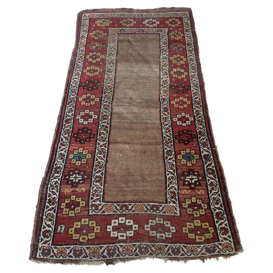 Full view of an antique Kurdish tribal wool rug from the 1870s featuring an open undecorated field in warm natural undyed camel wool, framed by a wide deep crimson border with bold geometric medallions including hooked diamonds and eight-pointed star rosettes.

