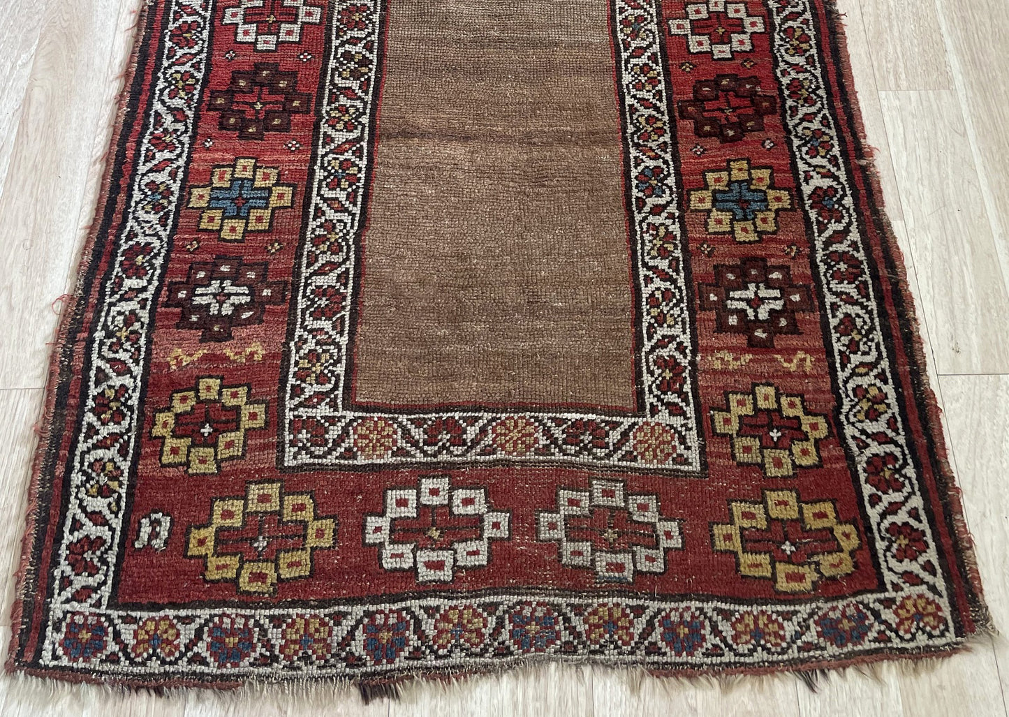 Close-up of a 150-year-old hand-knotted Kurdish rug showing the rich camel/tobacco field, a crimson main border with varied geometric motifs in ivory, saffron gold, dark brown, and teal blue, plus crisp ivory guard borders with vine-and-leaf scrolls.

