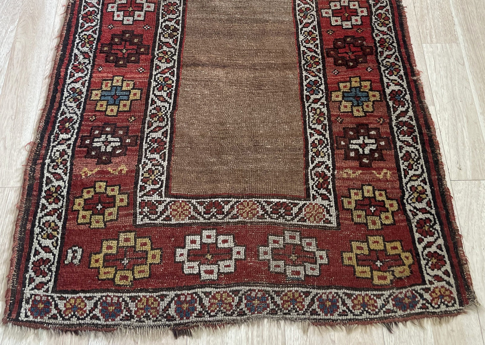 Close-up of a 150-year-old hand-knotted Kurdish rug showing the rich camel/tobacco field, a crimson main border with varied geometric motifs in ivory, saffron gold, dark brown, and teal blue, plus crisp ivory guard borders with vine-and-leaf scrolls.

