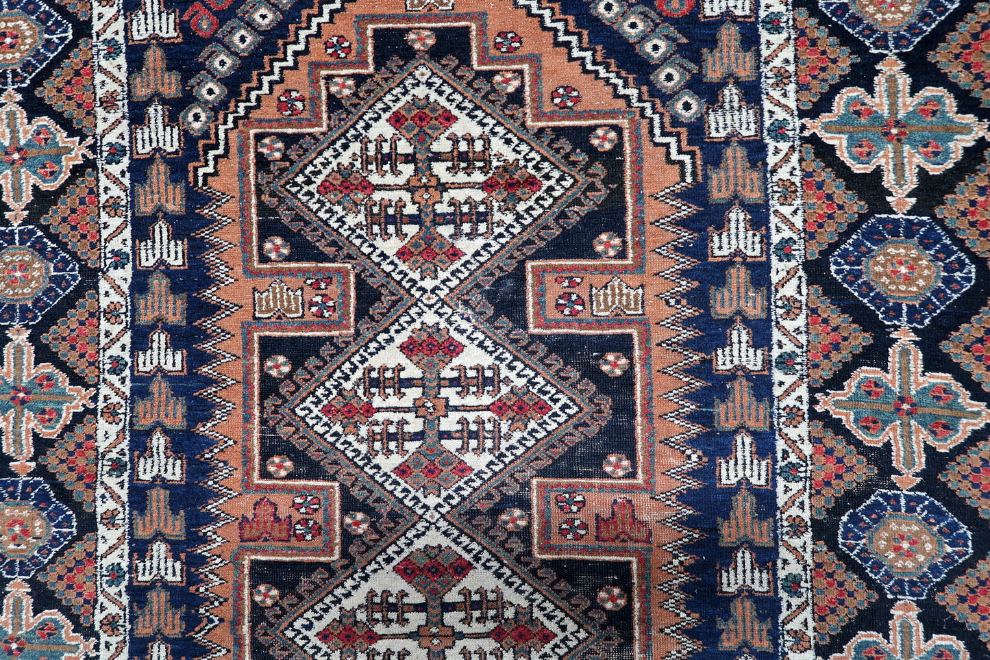 Close-up of an antique Persian tribal rug's central design, highlighting the detailed diamond medallions and geometric motifs in rich, warm colors.

