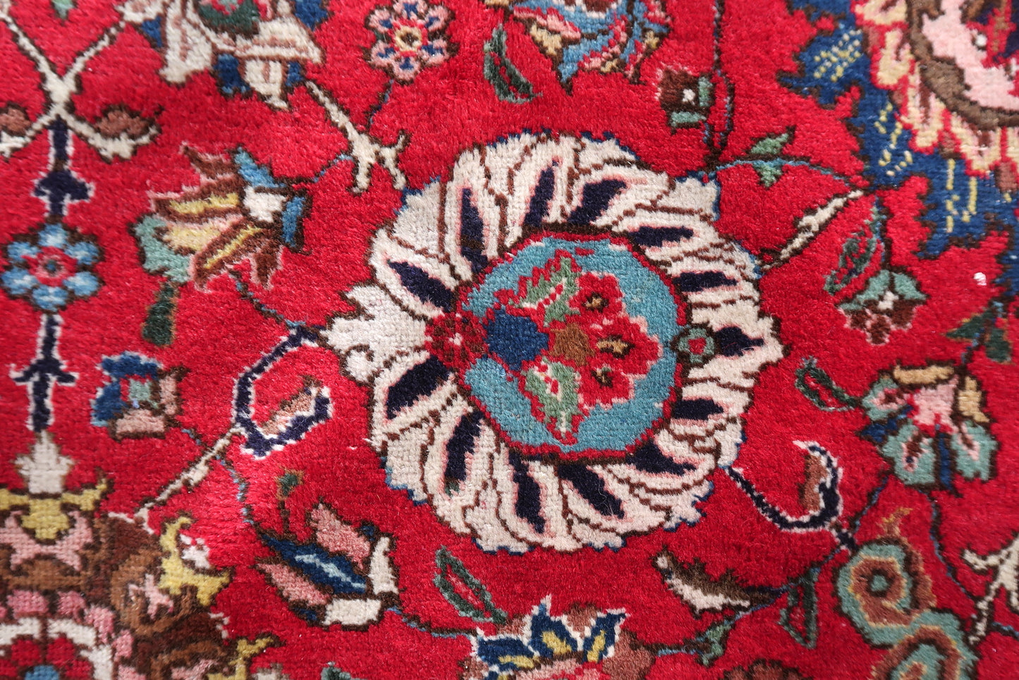 Close-up of the corner of a 1920s Tabriz rug, revealing the transition from the crimson allover floral field to the ivory main border with red palmettes and the teal guard border with rosette vine scroll.

