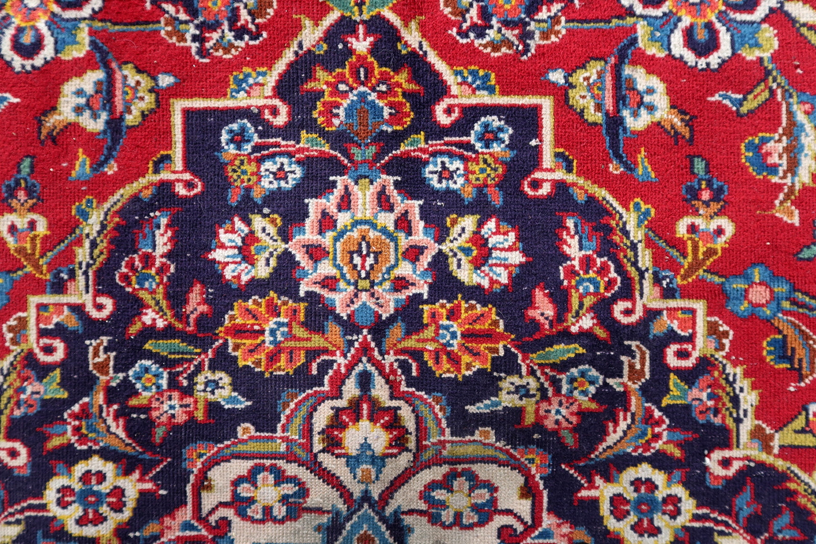 Close-up of the corner of a 1920s Kashan rug, revealing the transition from the deep crimson allover floral field to the navy spandrel, and the multi-layer border system with sky blue and ivory guards.

