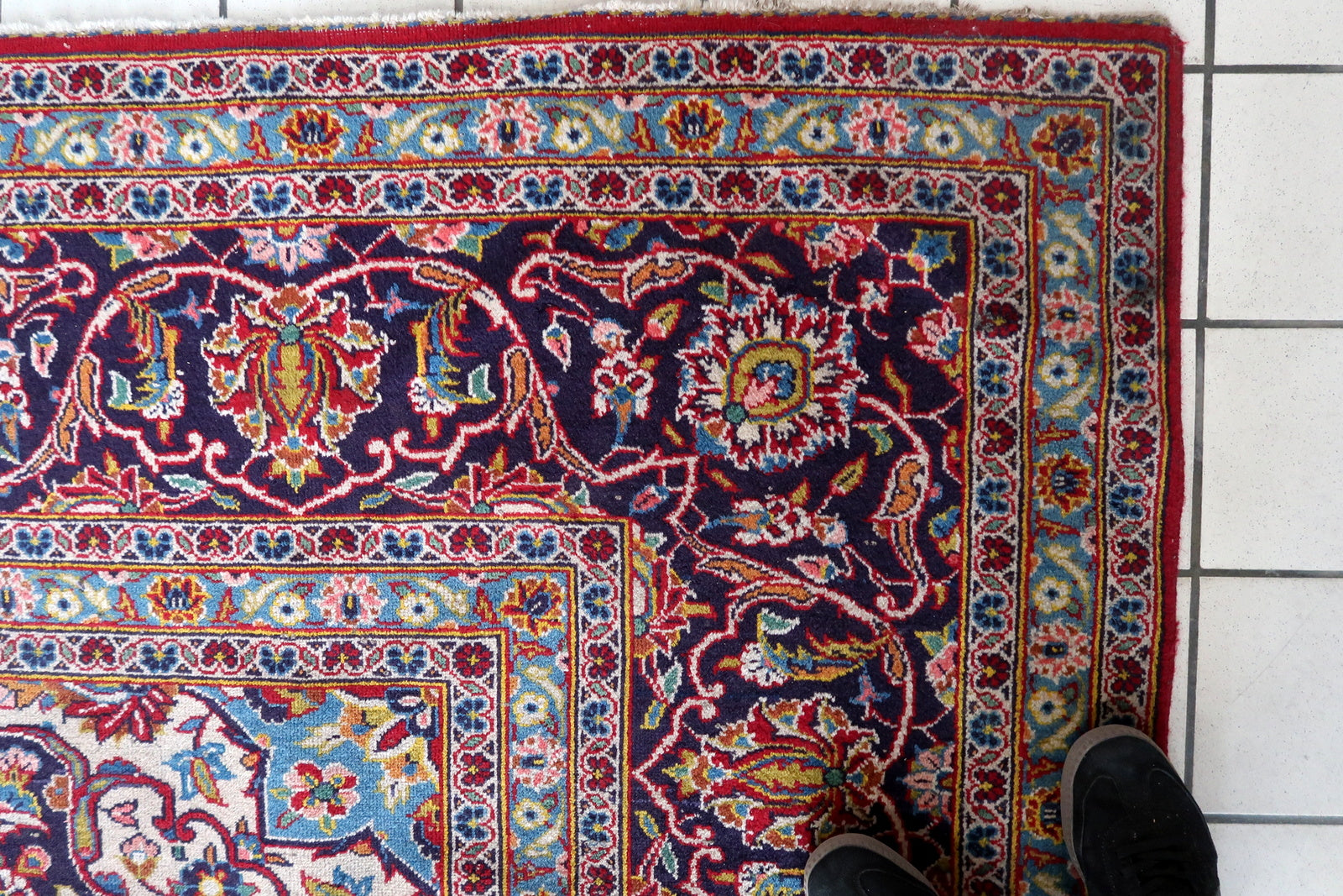Close-up of the central medallion on a 1920s Persian Kashan carpet, showing a deep navy/indigo multi-lobed form with an ivory center enclosing a sky blue cross-star with internal floral detail in red, pink, and gold, surrounded by a pendant outline with pointed extensions.

