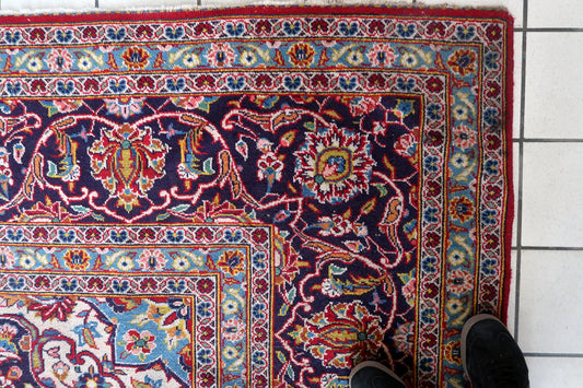 Close-up of the central medallion on a 1920s Persian Kashan carpet, showing a deep navy/indigo multi-lobed form with an ivory center enclosing a sky blue cross-star with internal floral detail in red, pink, and gold, surrounded by a pendant outline with pointed extensions.

