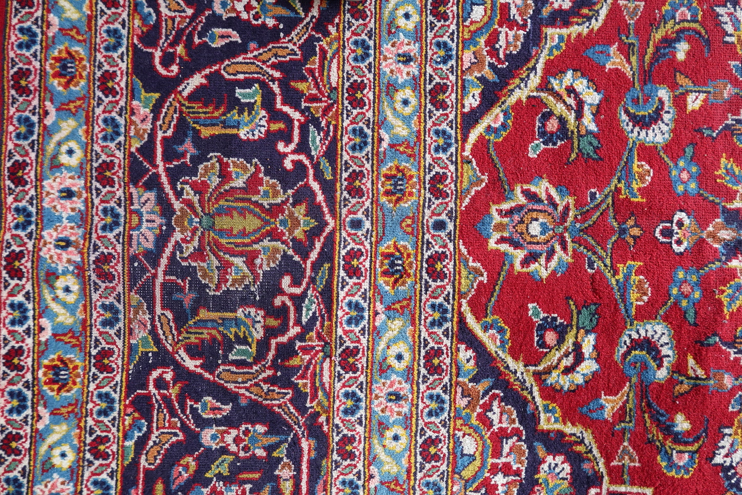 Detail of the border system on a vintage Persian Kashan rug, showing a deep navy main border with ivory arabesque scroll, vivid sky blue inner guard borders with floral rosette vine scroll, and ivory outer guard borders with small floral meander.

