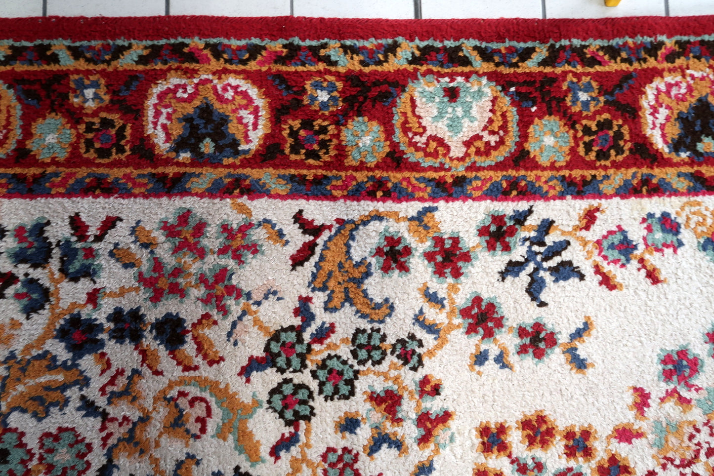 Detail of the border system on a vintage French Savonnerie carpet, showing a wide deep crimson main border with large ivory floral medallions (roses and peonies with teal and gold detail) connected by a crimson vine scroll, framed by a teal inner guard border with geometric meander and a dark navy outer guard with gold meander.

