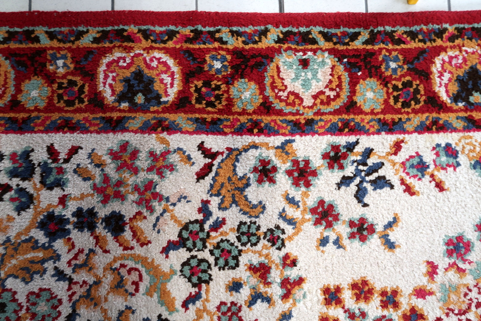 Detail of the border system on a vintage French Savonnerie carpet, showing a wide deep crimson main border with large ivory floral medallions (roses and peonies with teal and gold detail) connected by a crimson vine scroll, framed by a teal inner guard border with geometric meander and a dark navy outer guard with gold meander.

