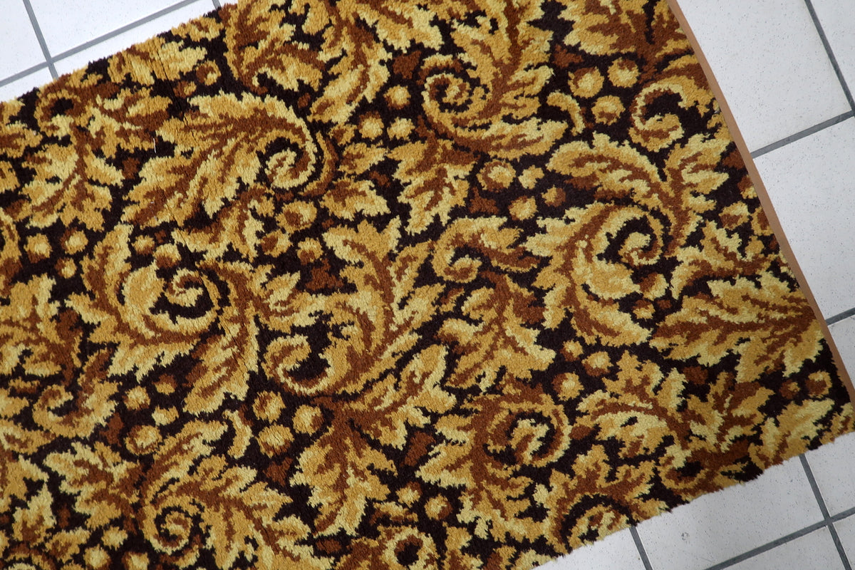 A close-up of a plush area rug with a classic damask pattern in golden yellow, brown, and black, laid out on a white tiled floor.
