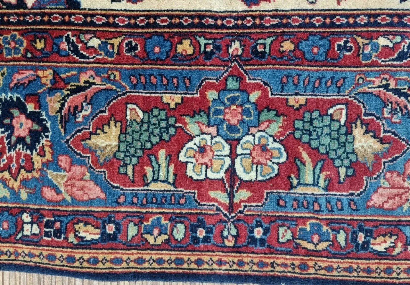 Detail of the border on a 1920s Persian Kashan carpet, revealing repeated floral and vine motifs in beige, navy, and cream with elegant symmetry and fine wool texture.


