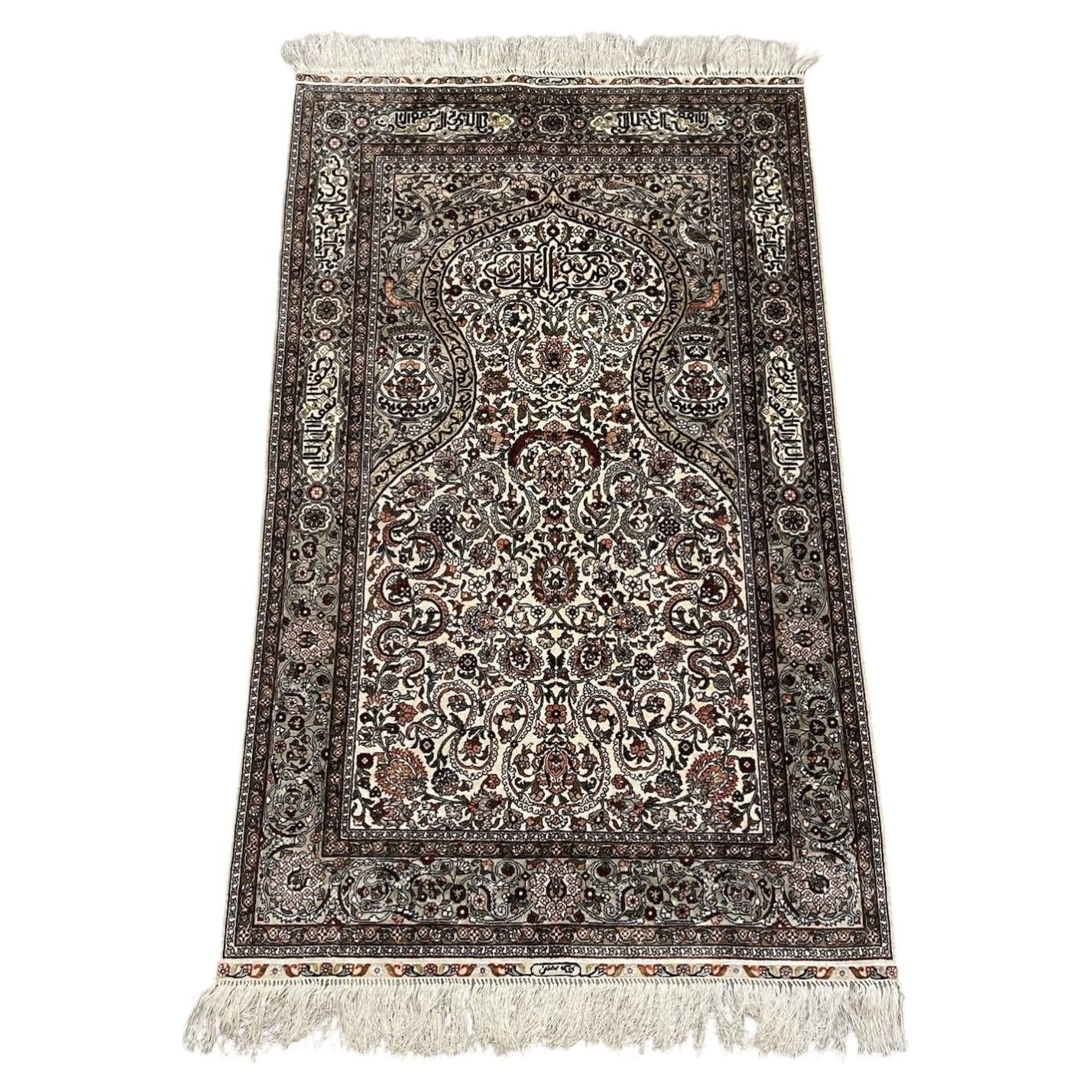 Full view of a vintage Turkish Hereke silk prayer rug from the 1970s, featuring a pointed mihrab arch on an ivory ground with Arabic calligraphy "Allahu Akbar", a paradise bird (simurgh/phoenix) above a double-gourd vase, and an olive-green arabesque border.

