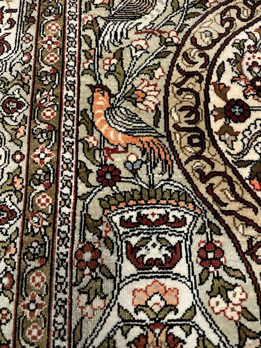 Close-up of a hand-knotted Hereke silk prayer rug with extraordinary knot density of 400–520 knots per square inch, showing a phoenix with orange-amber body and blue-green wing feathers on an ivory silk field.

