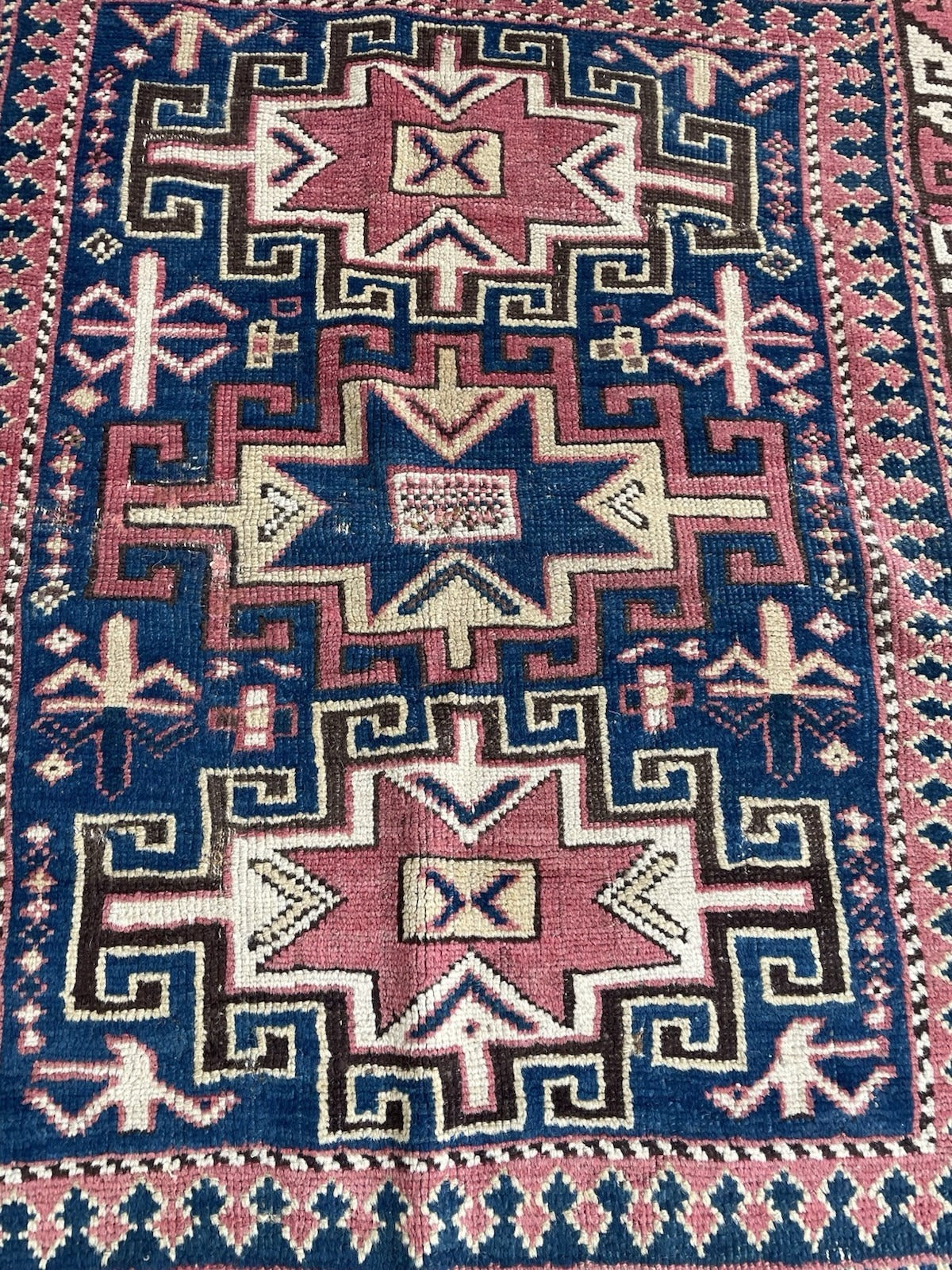 Antique Caucasian geometric rug with a large stepped shield medallion in dusty rose on royal blue, the medallion interior containing a dark brown field with an ivory sunburst or stepped cross motif.

