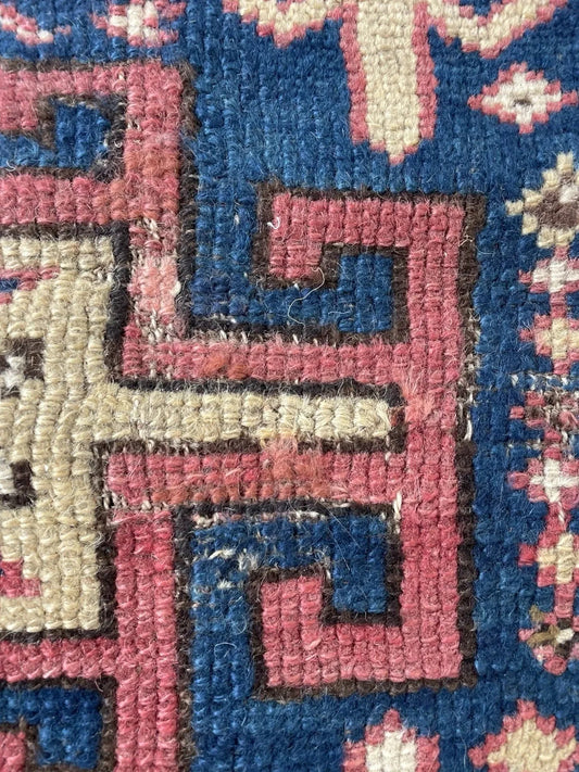 Close-up of a 1880s Kazak rug showing the central medallion interior: a dark brown field containing a large ivory stepped cross with four projecting arms terminating in stepped hooks, and a small ivory rectangle with a blue cross protective amulet.

