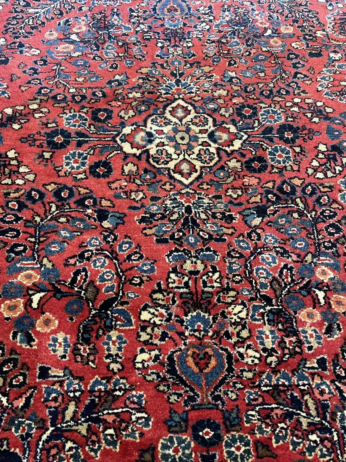 Close-up of the corner and border detail on an antique Sarouk wool rug, showing large palmette blossoms and curling arabesques in crimson, ivory, and rose.

