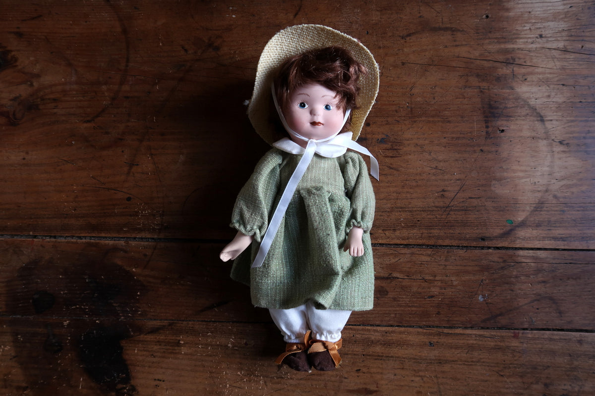 Porcelain doll face with blue-grey eyes, short brown hair and white chin ribbons.