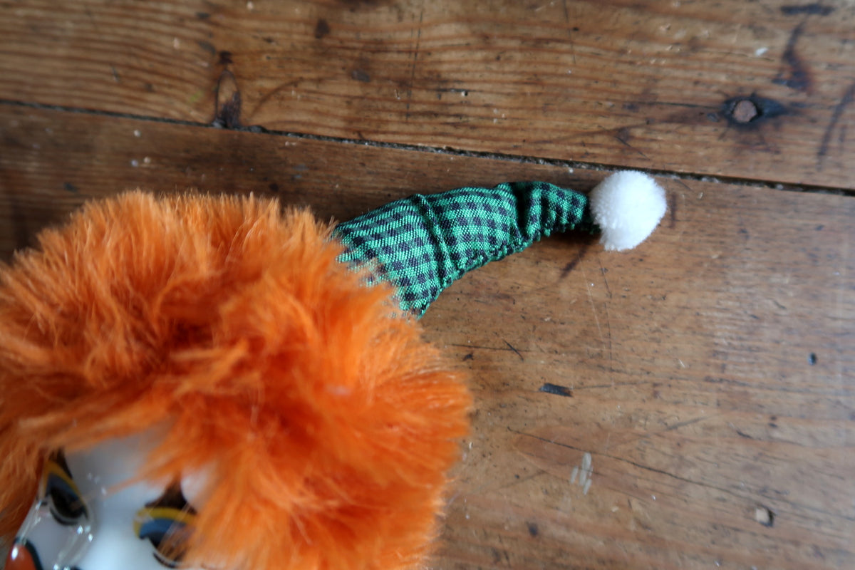 Green gingham pointed hat with a white pompom above tousled orange hair.