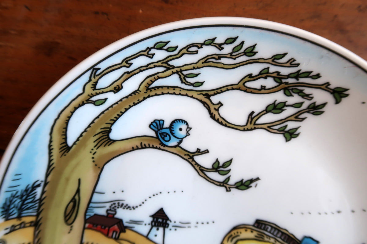 Blue bird perched on a leafy tree branch above the coastal scene on the Sommer plate.