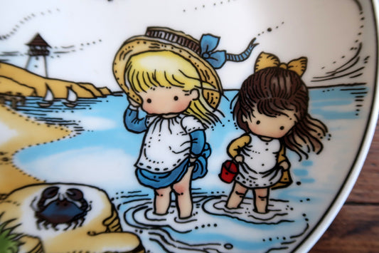 Two girls standing barefoot in shallow water, one with a straw hat and blue ribbon, the other with a yellow bow.