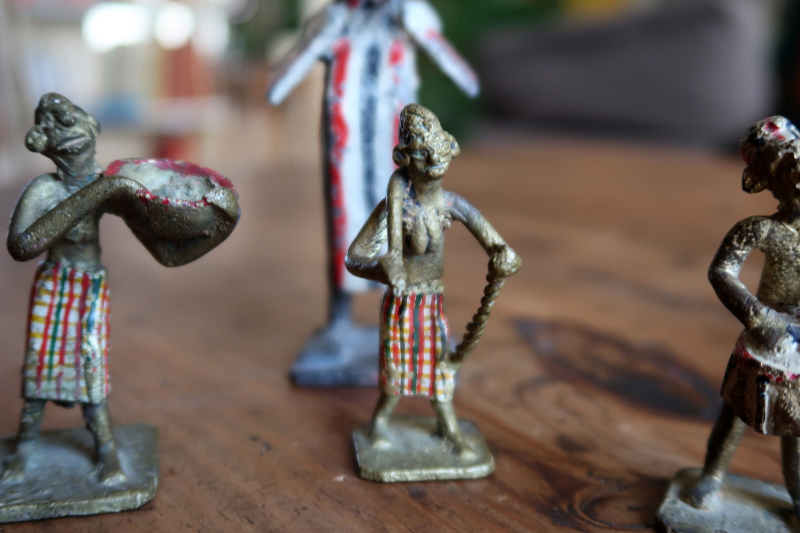 Detail of three small attendant figurines surrounding a central ceremonial figure, showing colorful striped loincloths in red, white, and green, with one holding a tam-tam drum, one carrying a staff or spear, and one in a composed upright pose.

